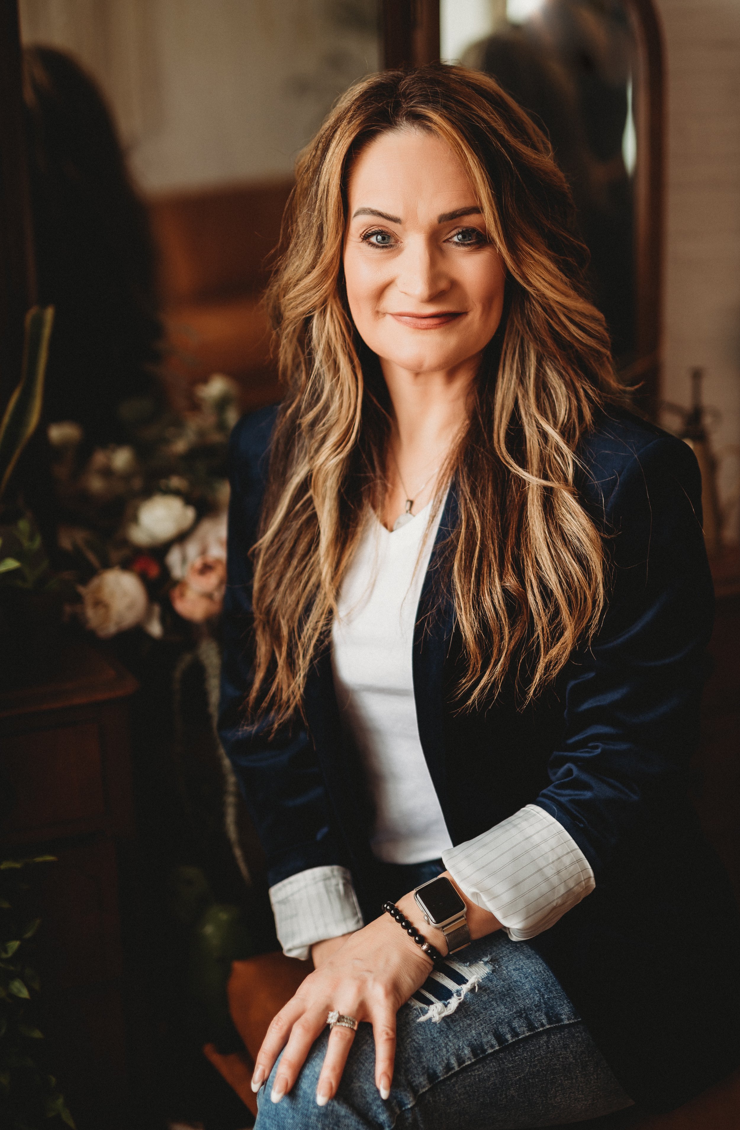 A woman with long wavy hair sitting confidently, wearing a navy blazer, white shirt, and jeans, accessorized with a smartwatch and bracelets. There are flowers and a mirror in the background.