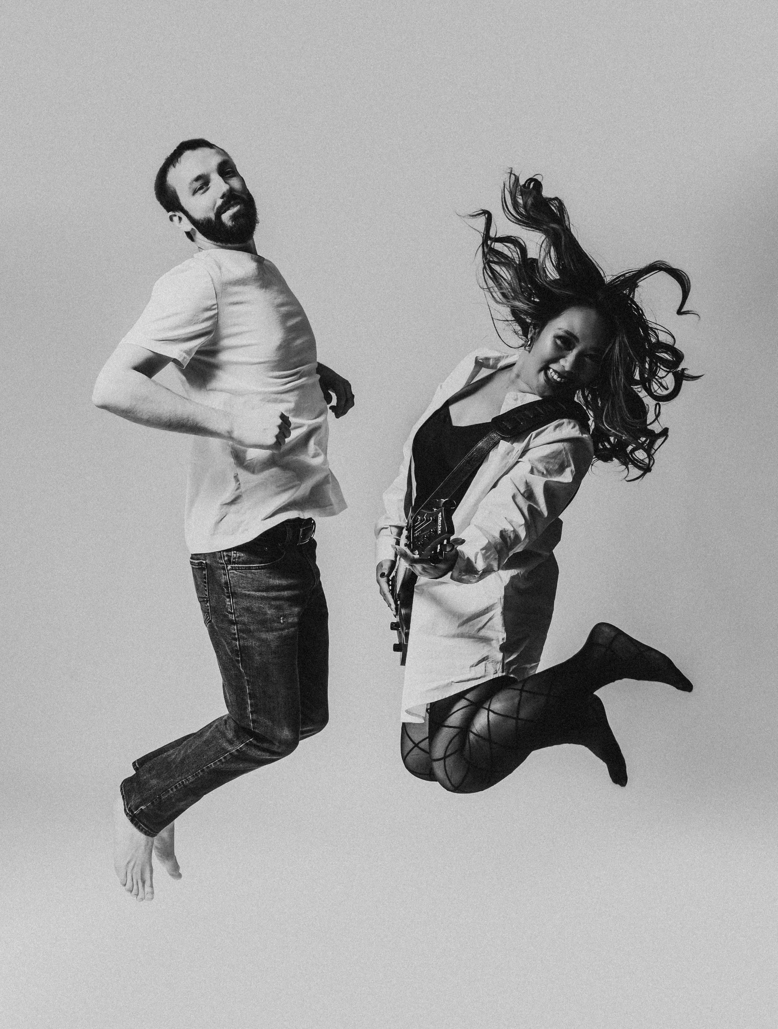 Black and white photo of a man and woman jumping, with the woman holding a guitar, showcasing dynamic movement.