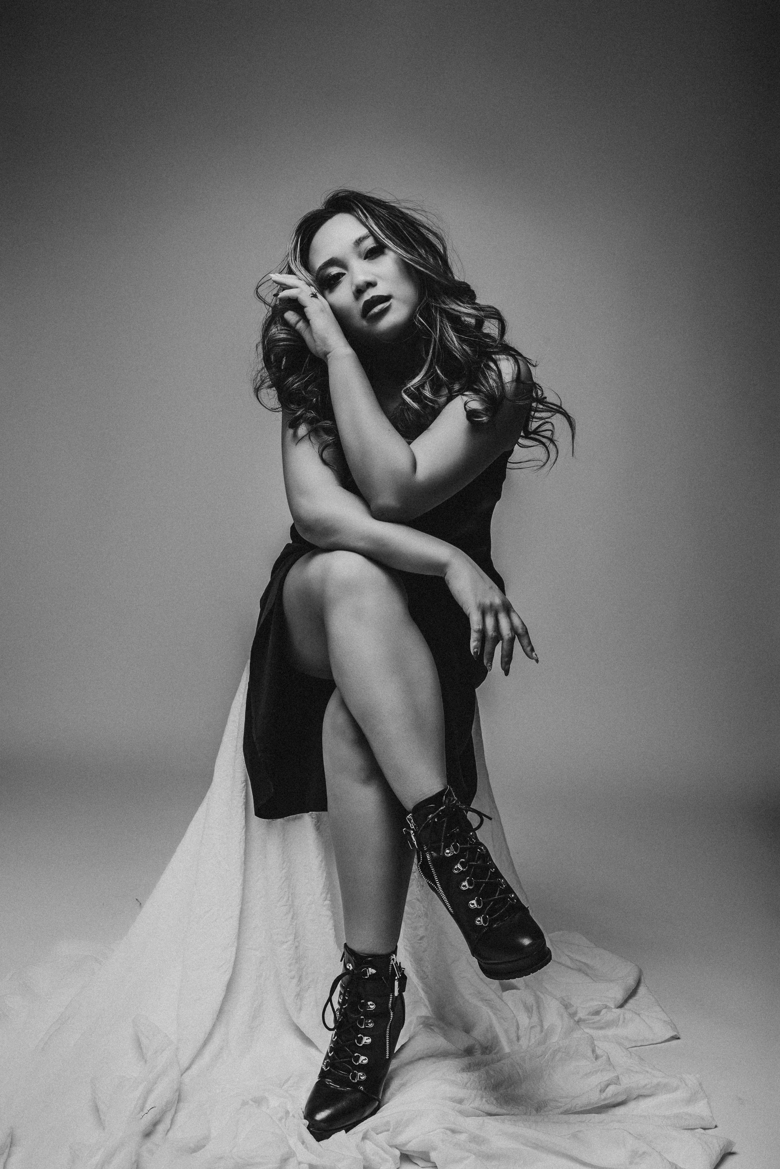 Black and white photo of a seated woman with curly hair, wearing a sleeveless dress and lace-up boots, posing thoughtfully.