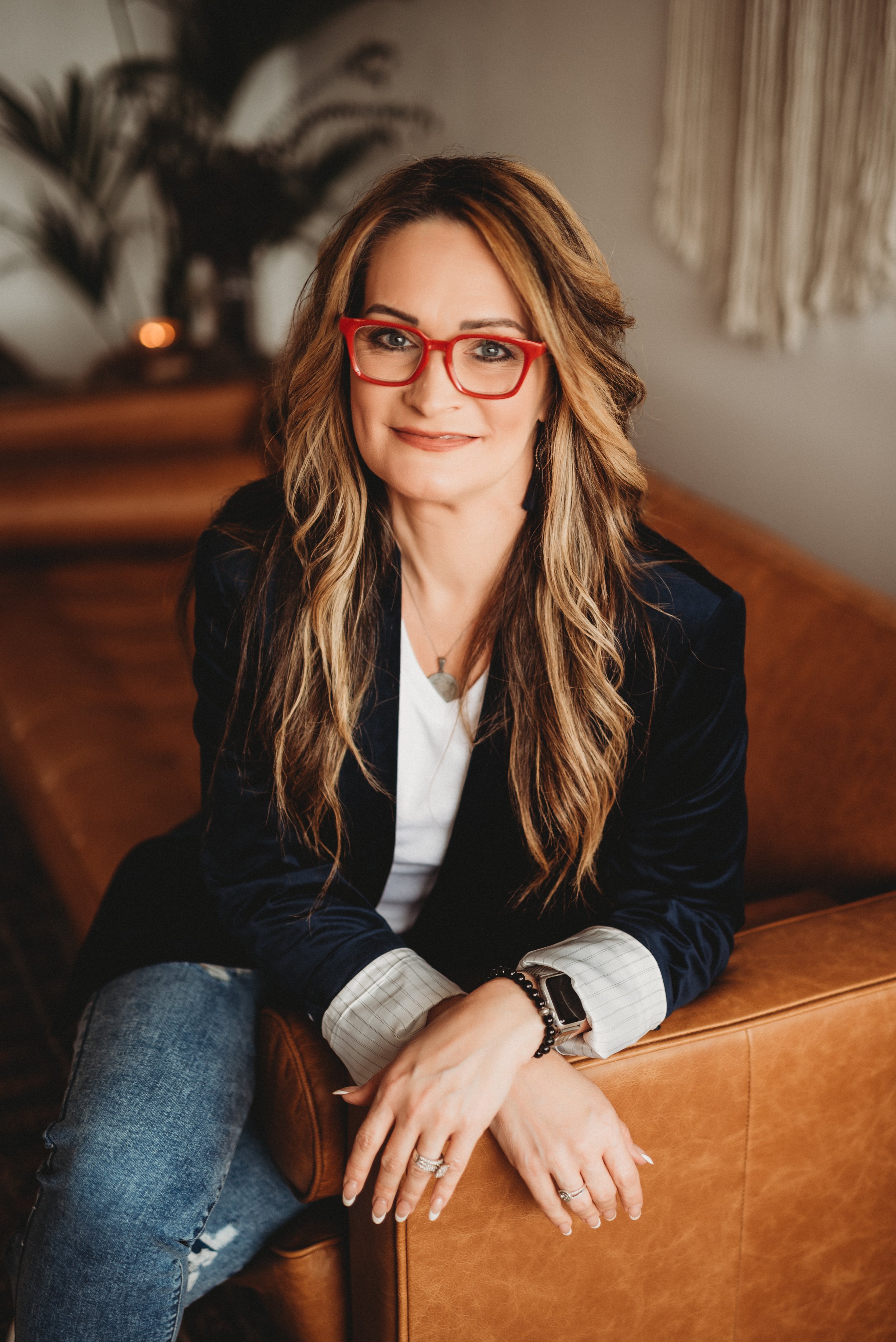 Woman with long hair and red glasses sitting on a brown couch, dressed in a dark jacket and jeans, in a cozy indoor setting.