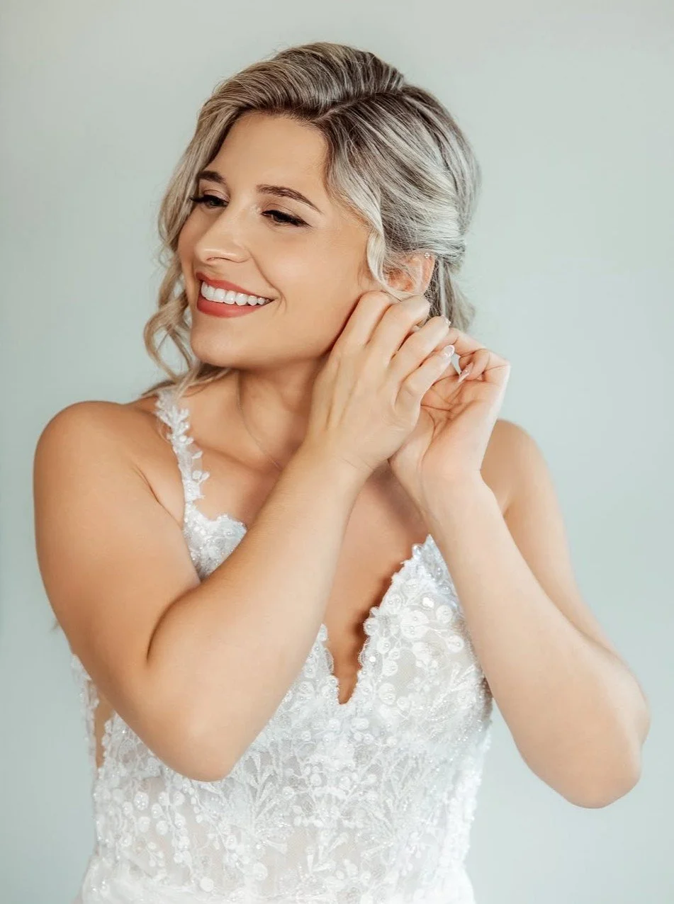A woman with grayish blonde hair, smiling, wearing a white lace dress, placing her earring in her ear.