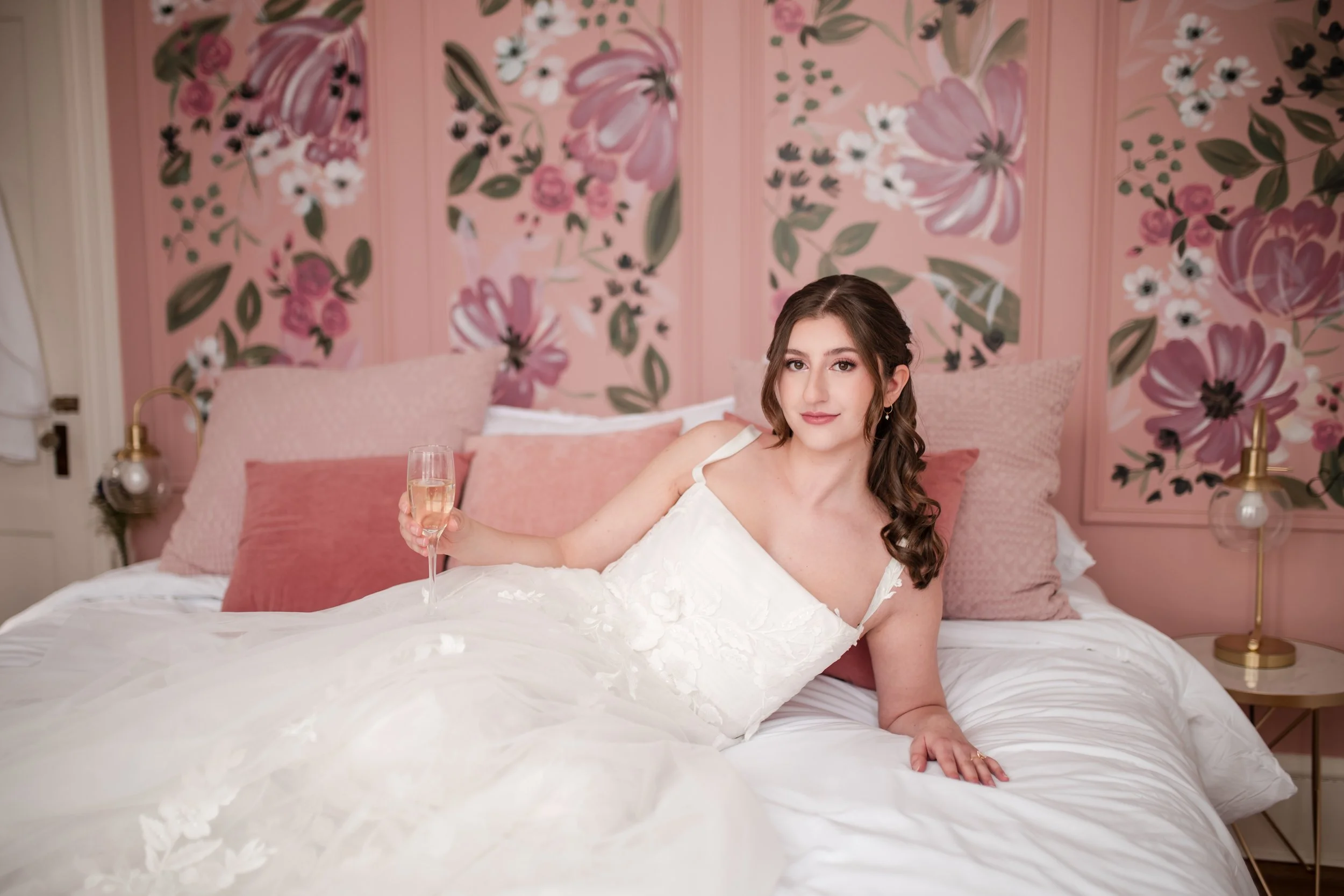 A young woman in a white dress lying on a bed with pink pillows, holding a glass of champagne, in a pink floral-themed room