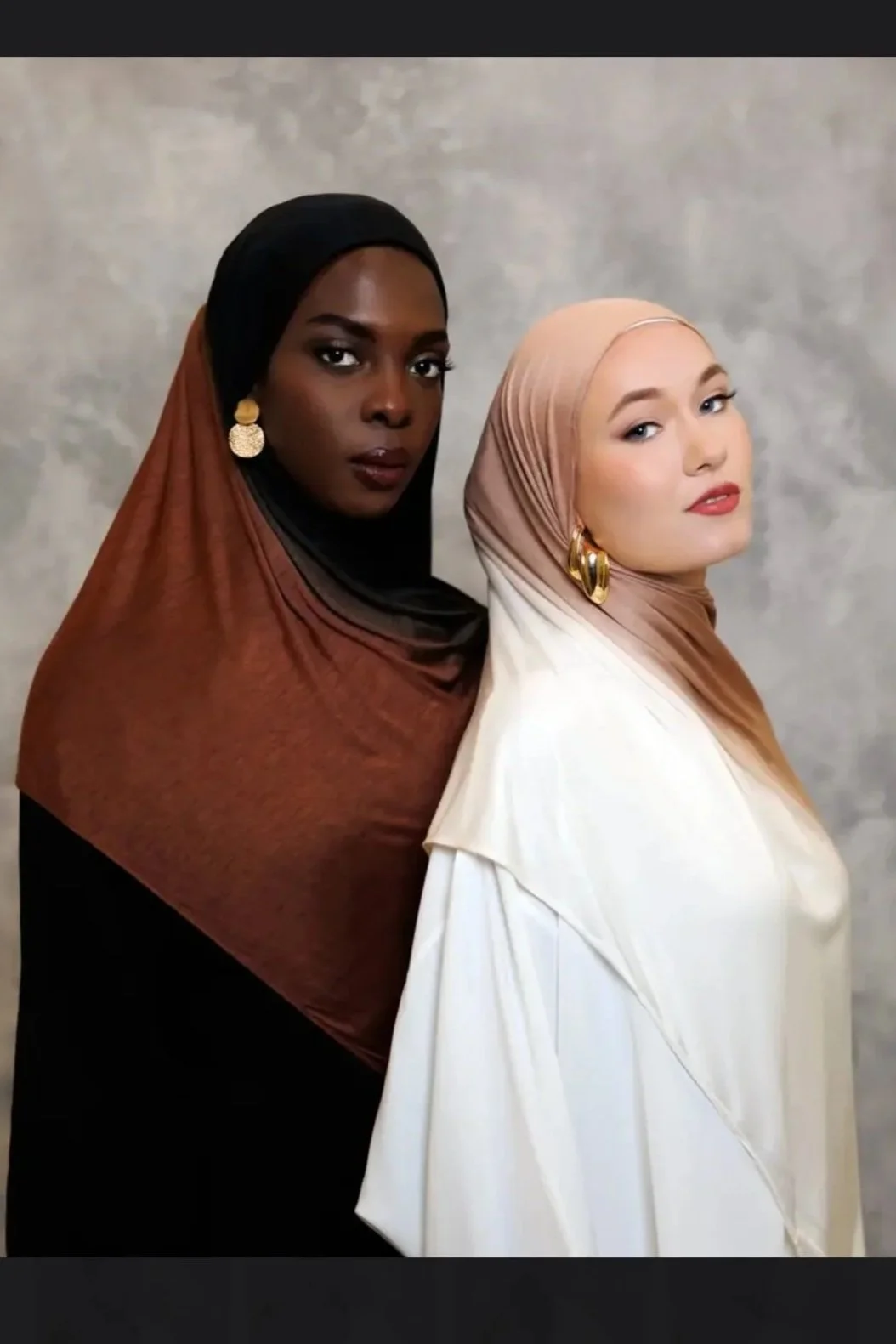 Two women wearing headscarves and elegant clothing, standing close together against a plain, neutral background. The woman on the left has dark skin, a black and brown headscarf, and gold earrings. The woman on the right has light skin, a beige headscarf, and gold earrings.