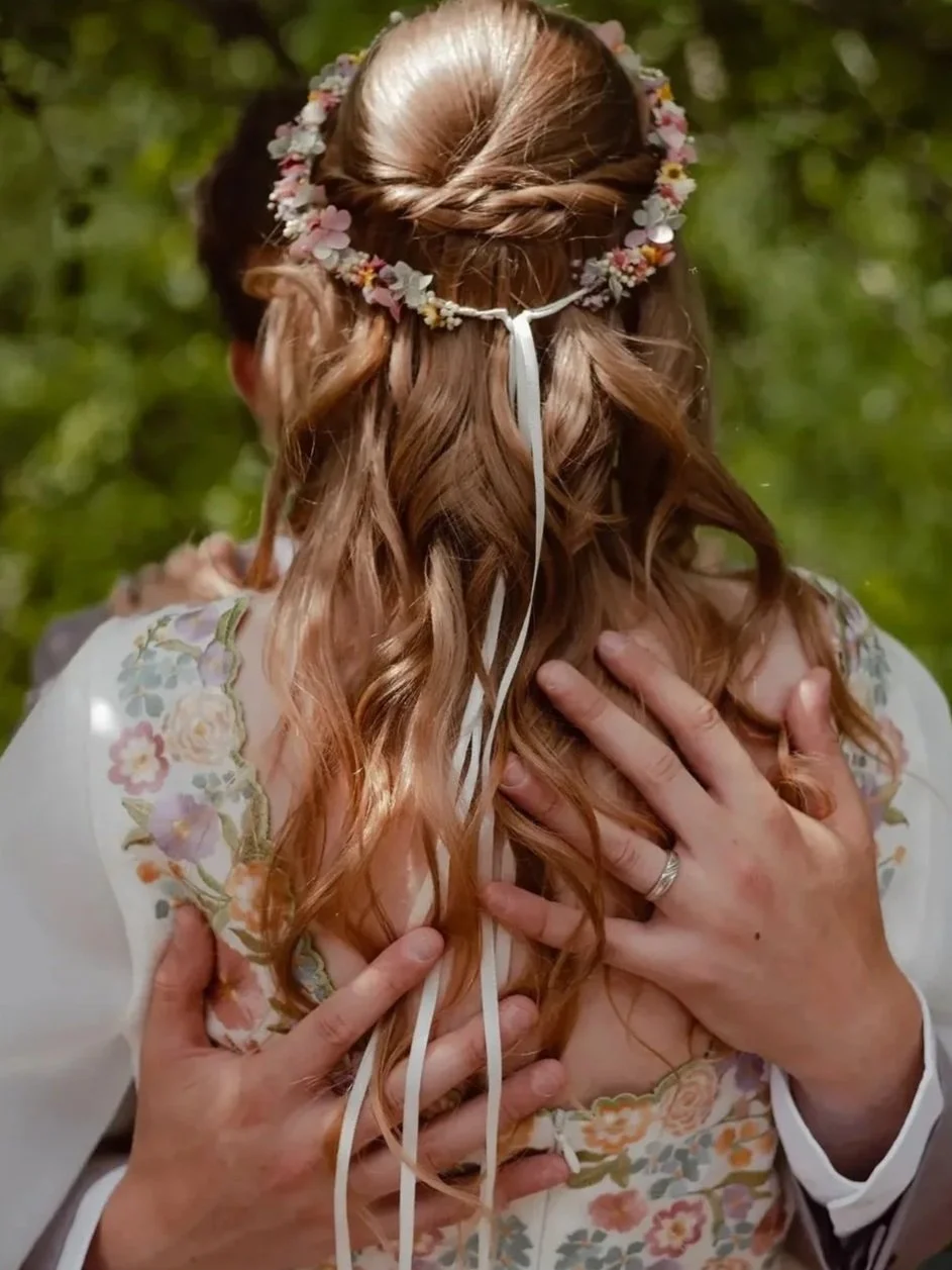 A woman with long, dark, wavy hair styled with loose curls, decorated with two pearl and floral hair accessories, sitting indoors on a gray chair, with a beige wall, a doorway, and a black piece of furniture in the background.