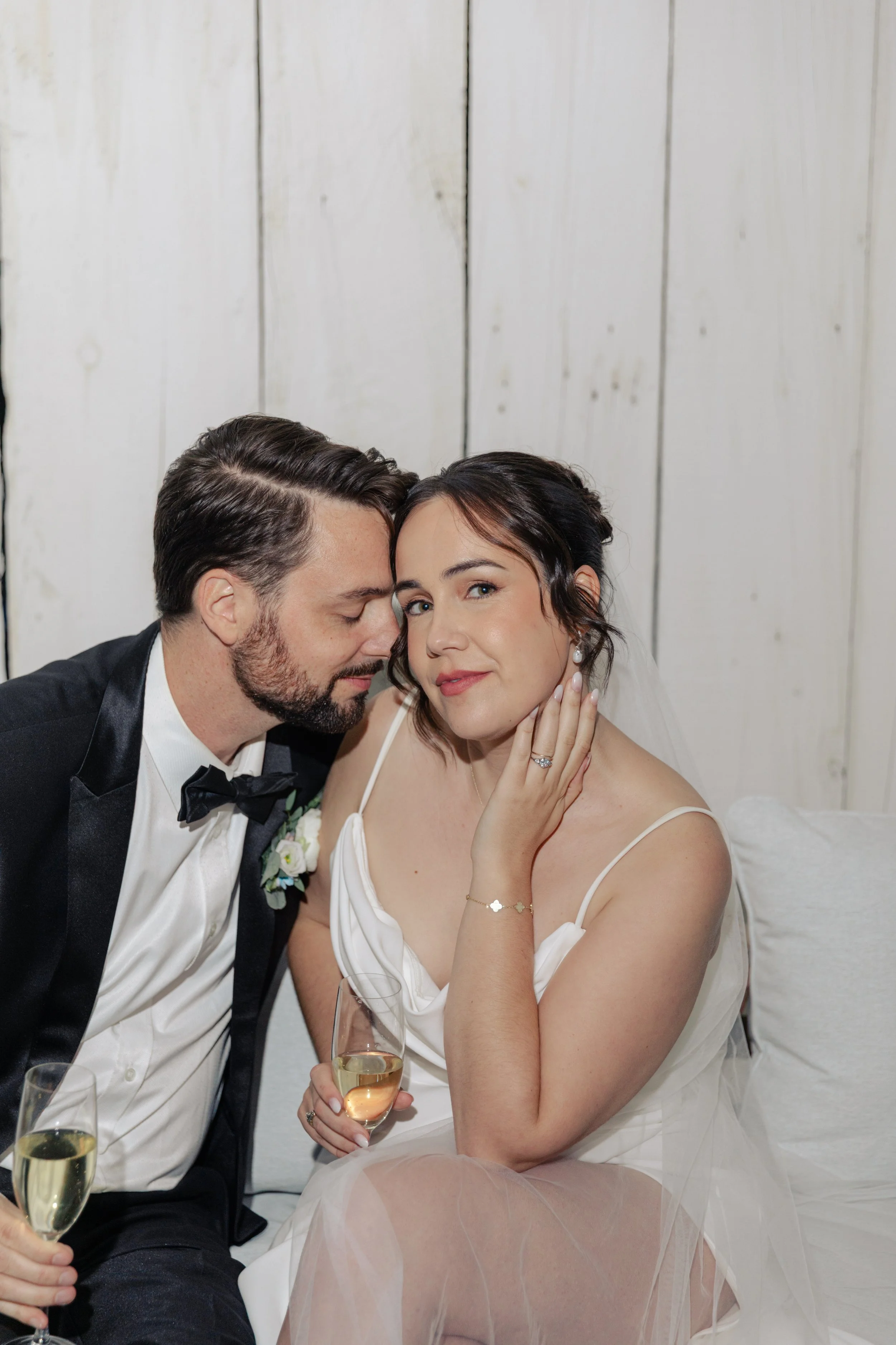 A bride and groom at their wedding, with the groom whispering in the bride's ear and the bride touching her face, holding a glass of champagne. They are dressed in wedding attire and seated at a celebration with a white wooden wall background.