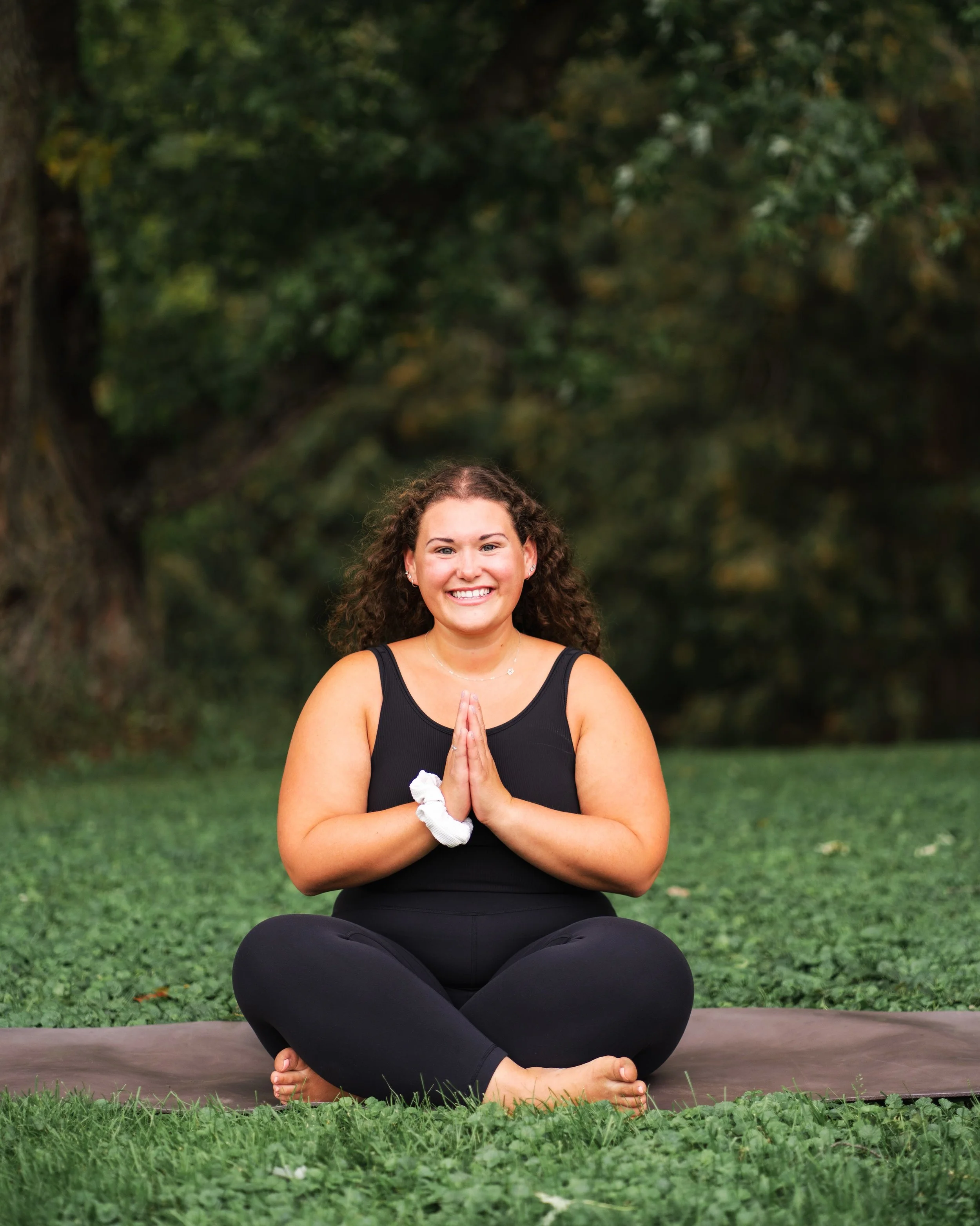 A woman with curly brown hair practicing yoga outdoors on a mat, sitting cross-legged with hands pressed together in prayer position, smiling at the camera.