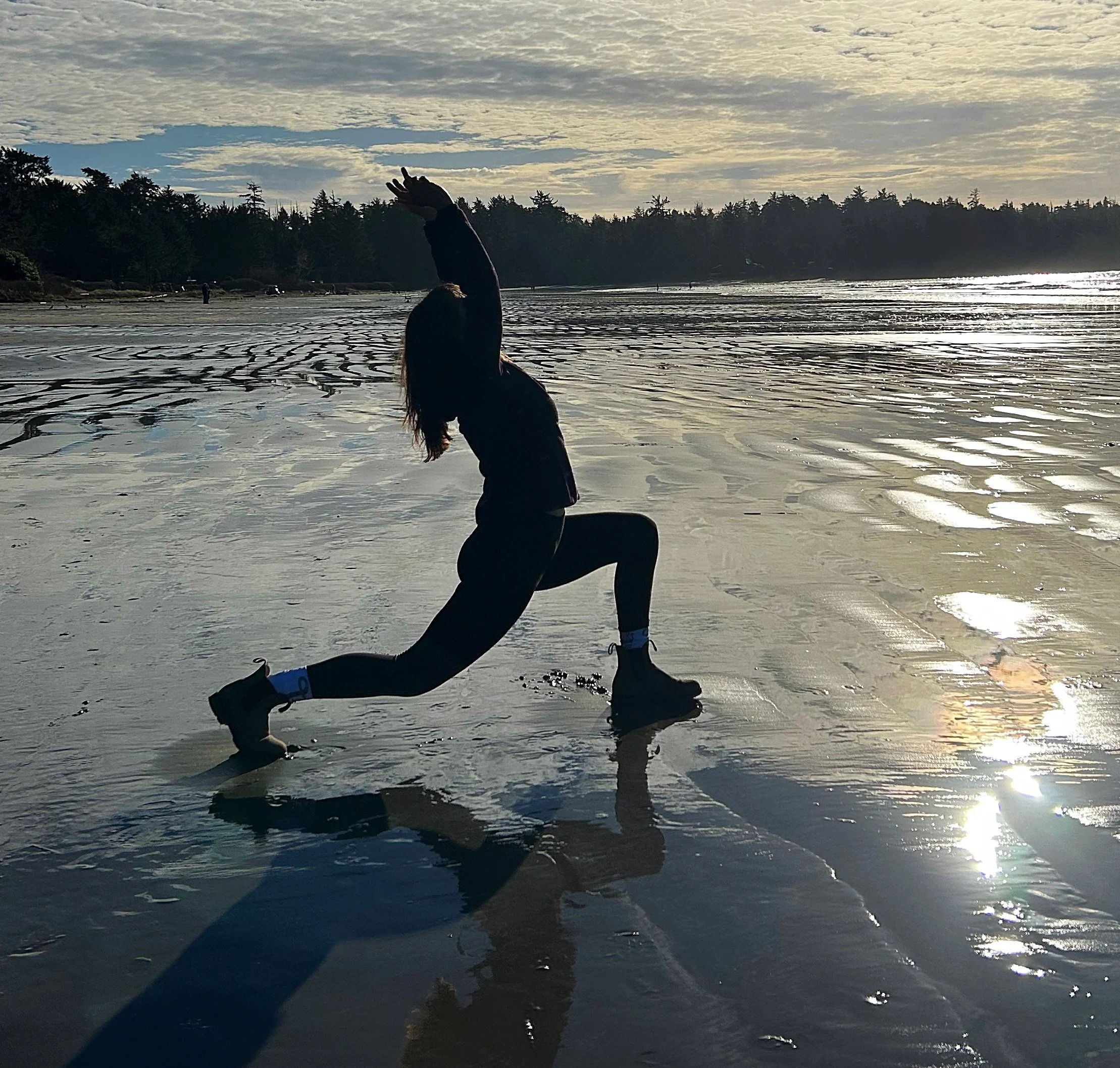 Person doing a yoga pose on a wet sandy beach during sunset or sunrise, with trees in the background.