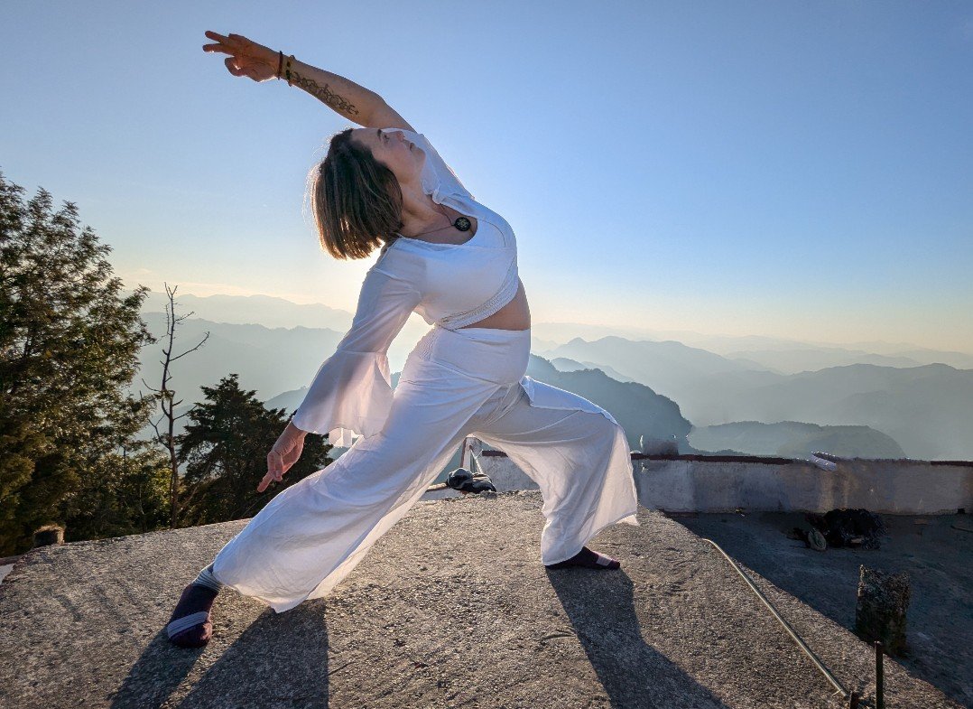 Woman practicing yoga on a mountain ledge at sunrise, wearing white flowing clothing, with scenic mountain ranges in the background.