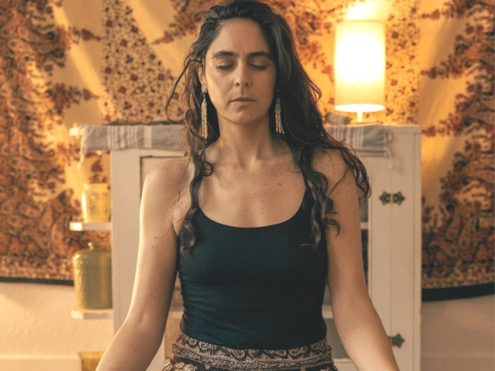A woman with long dark curly hair and wearing a black sleeveless top, sitting cross-legged with her eyes closed in a warmly lit room with patterned wallpaper and a white cabinet in the background.