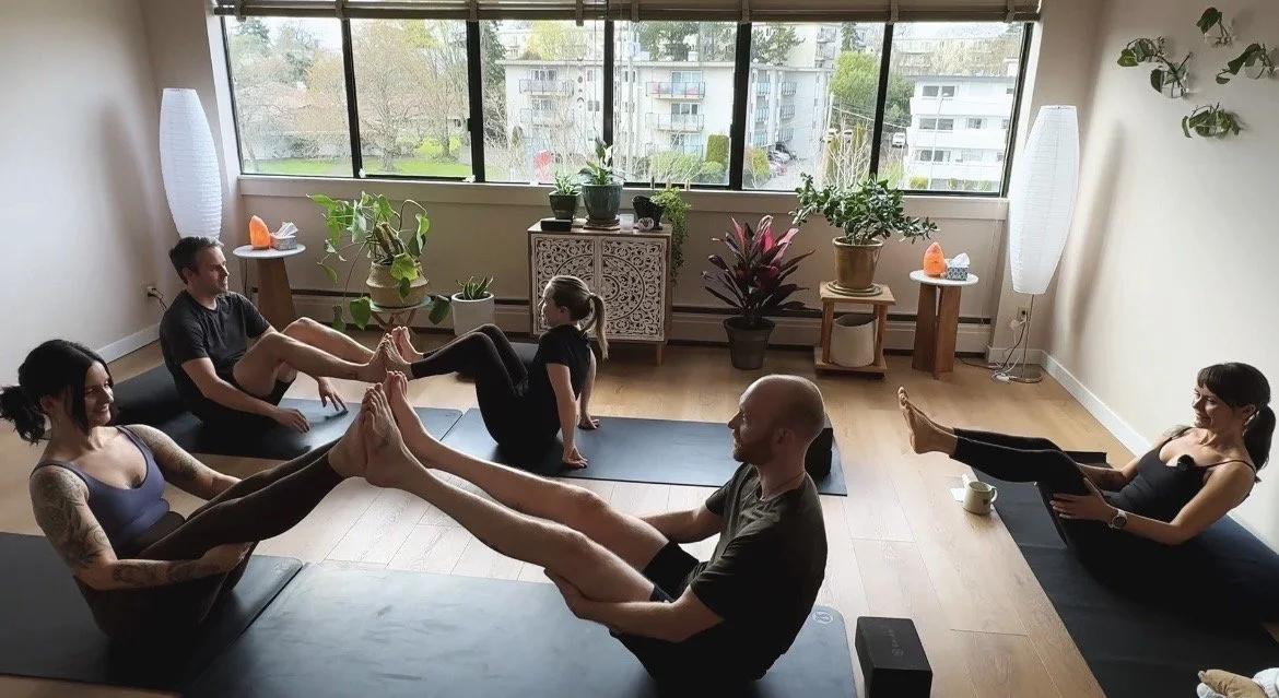 Five people participating in a yoga class in a bright room with large windows and indoor plants, sitting on mats in pairs doing partner stretches.