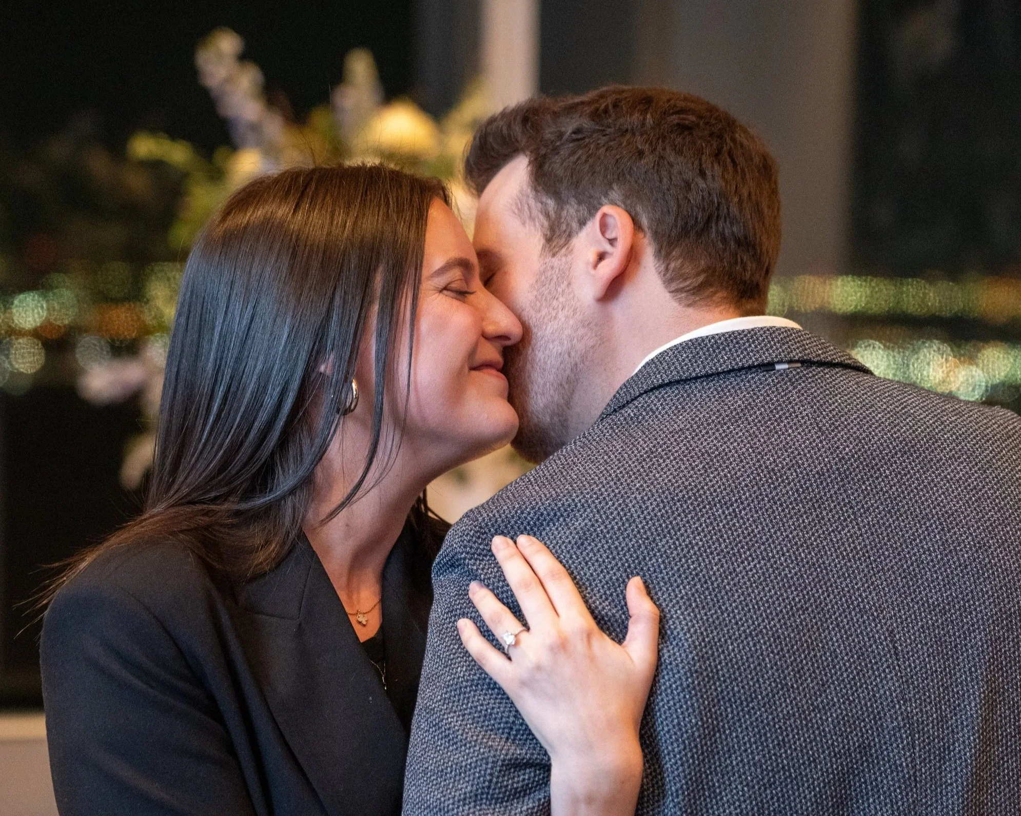 Groom whispers to fiancée on Brooklyn rooftop as her diamond ring sparkles with Manhattan skyline behind.