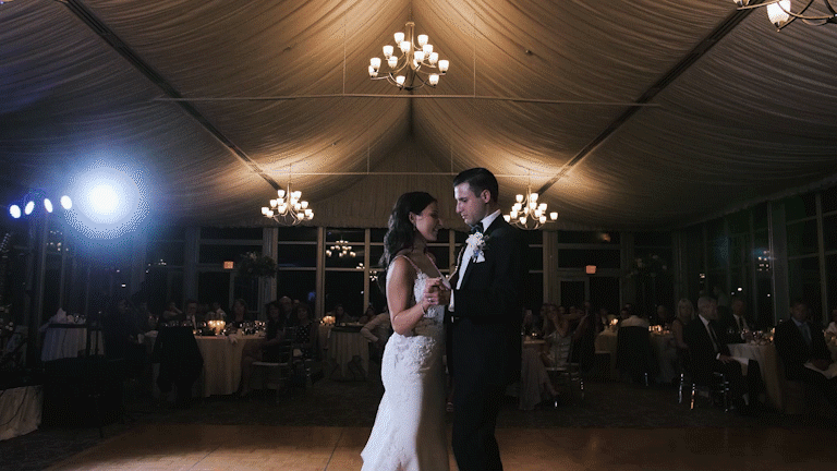 A bride and groom dancing in a dimly lit reception hall with chandeliers and guests seated at tables in the background.