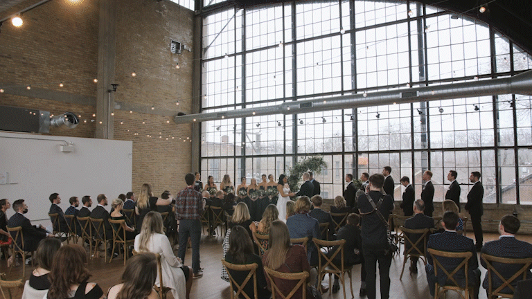 Indoor wedding ceremony with guests seated in chairs, bride and groom at the altar, and large industrial-style windows in the background.