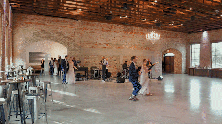 Couples dancing on a spacious dance floor with a live band playing music in a rustic venue with brick walls and large windows.