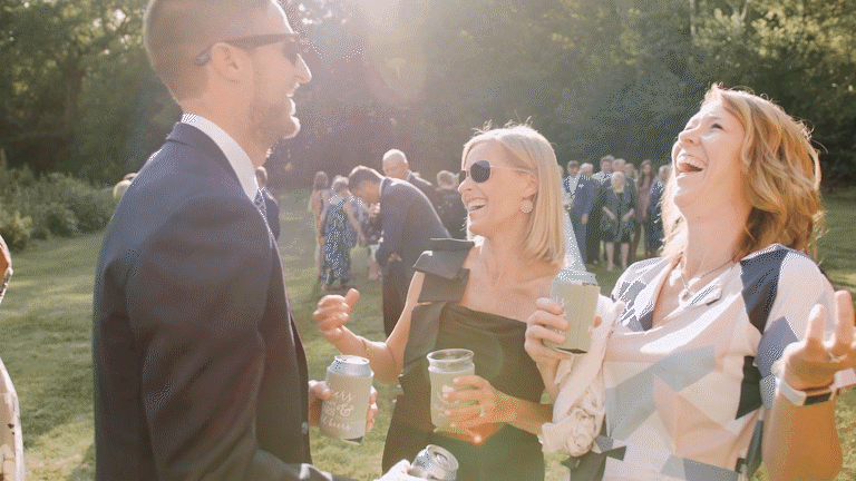 Three people, two women and one man, laughing and holding drinks at an outdoor gathering on a sunny day, with other people mingling in the background.