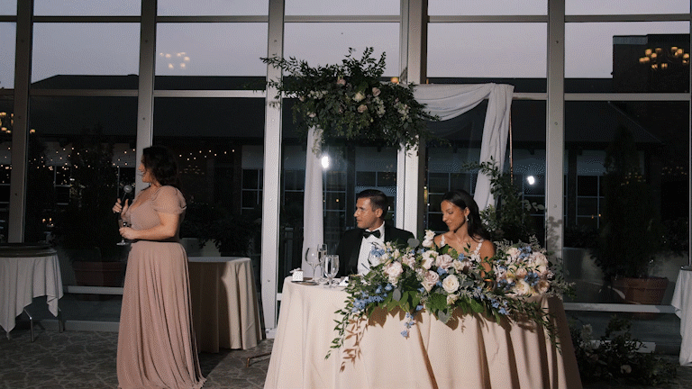 A bride and groom seated at a wedding reception table with floral decorations, a woman giving a speech, in a room with large windows and draped fabric decor.