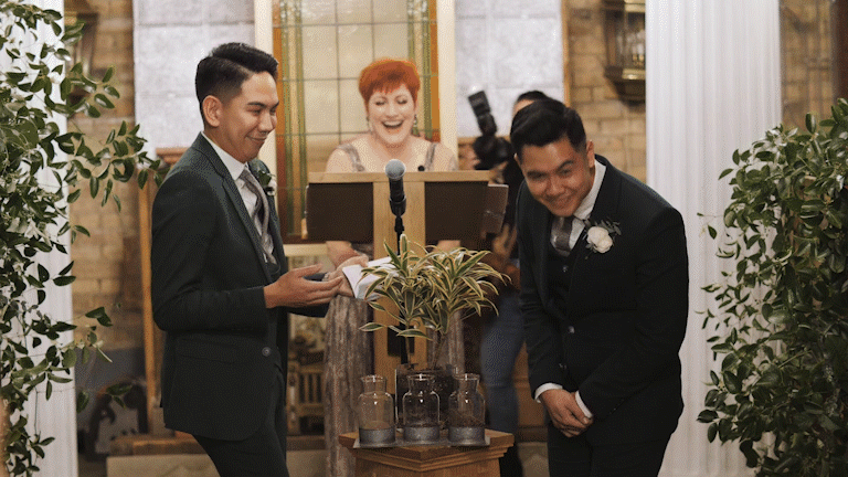 A wedding ceremony with two men in suits exchanging vows in front of an officiant, with a woman standing behind them, all smiling indoors decorated with plants.