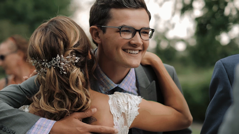 A man and woman sharing a hug at an outdoor wedding, with the woman wearing a lace dress and a hair accessory, and the man wearing glasses and a suit, both smiling.