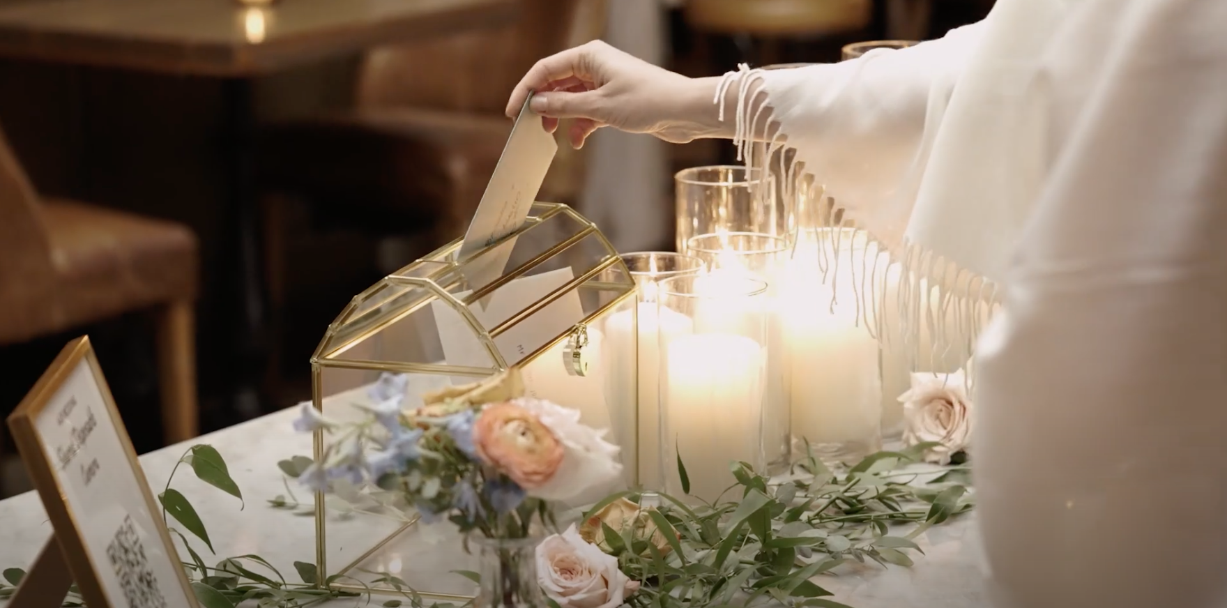 Person in a white fringed shawl placing a guest book into a gold geometric box on a wedding reception table decorated with white and pink flowers and lit candles.