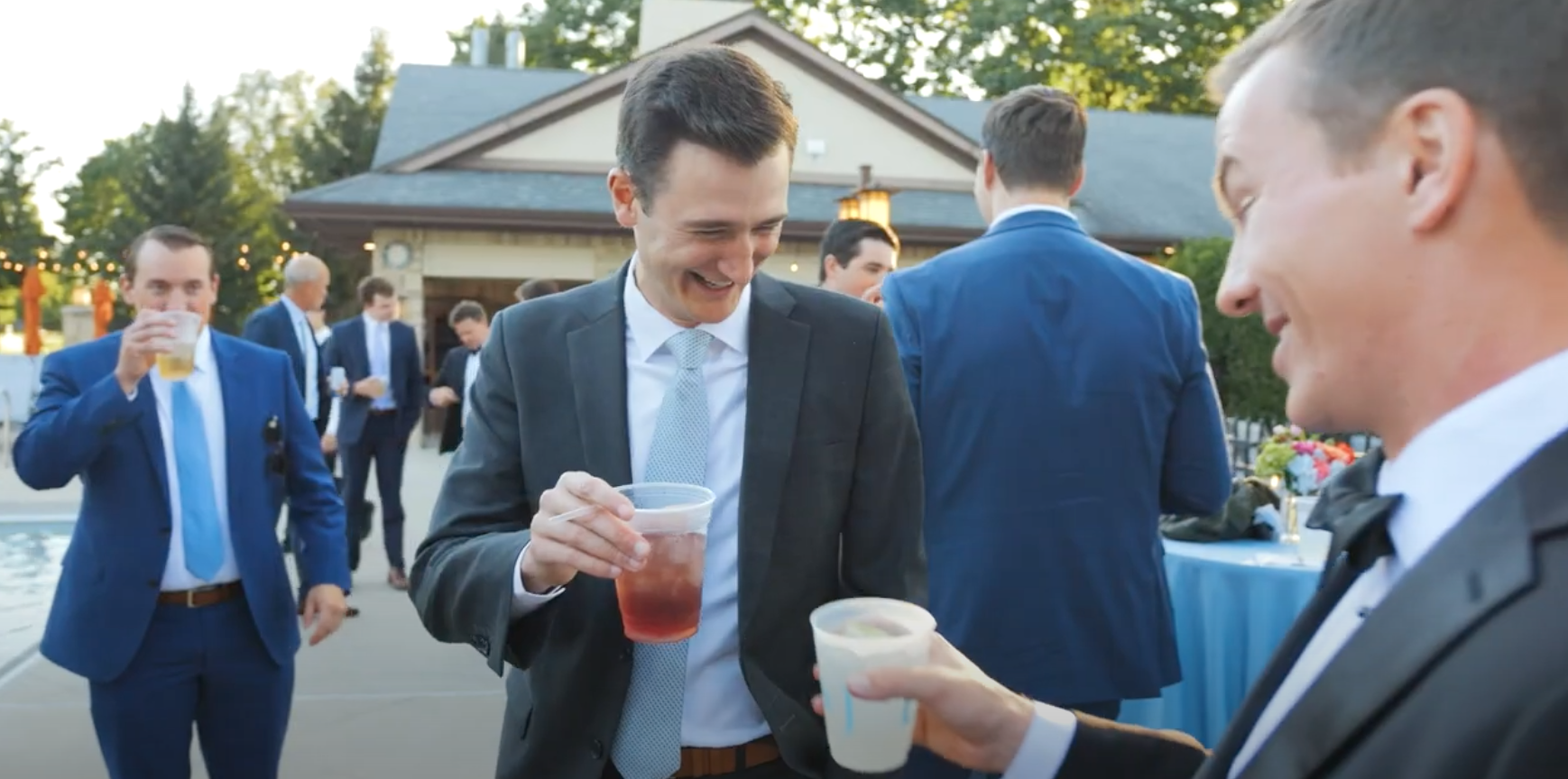 Men in suits at an outdoor gathering enjoying drinks and conversing, with a house and string lights in the background.