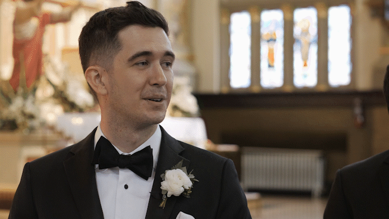 A man in a tuxedo with a white boutonniere, smiling during a wedding ceremony inside a church.