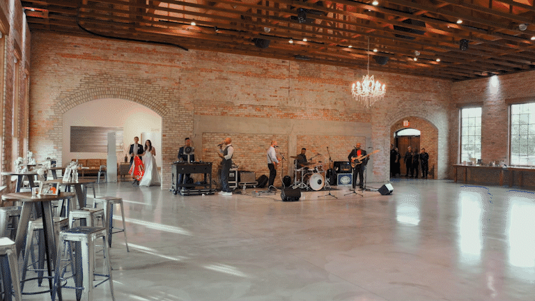 Band setting up music equipment in a spacious event hall with brick walls, large windows, and a chandelier, while a bride and groom dance nearby.