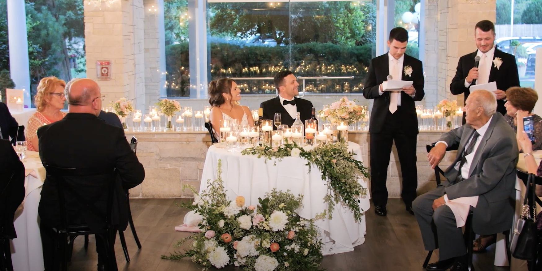 Wedding reception with bride and groom seated at head table, surrounded by family and friends, guests giving speeches, decorated with candles and floral arrangements.