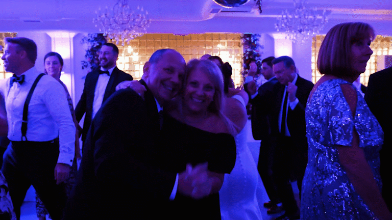 People dancing and socializing at a formal party or celebration in a decorated ballroom with chandeliers and festive lighting, including a smiling couple in the foreground.