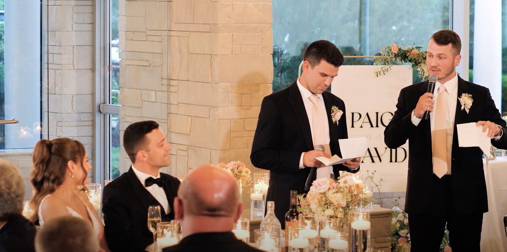 Groom and best man giving speech at wedding reception with bride and guests listening, decorated with candles and flowers.