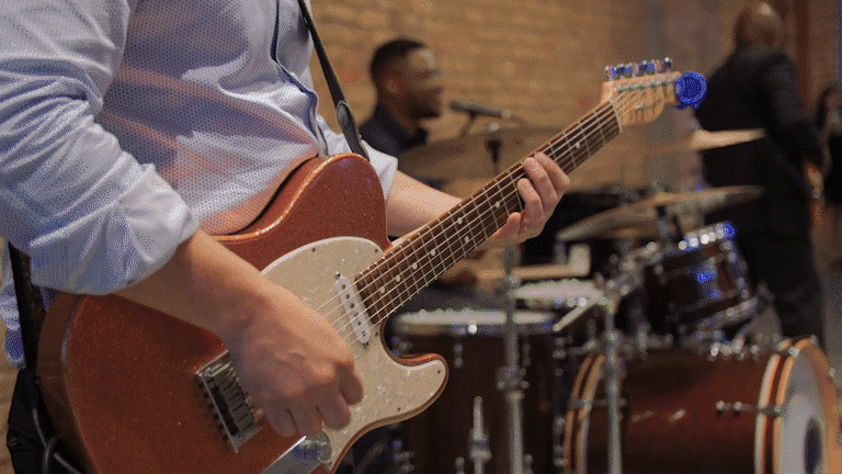 Person playing an electric guitar during a musical performance in an indoor venue with drums and other musicians in the background.