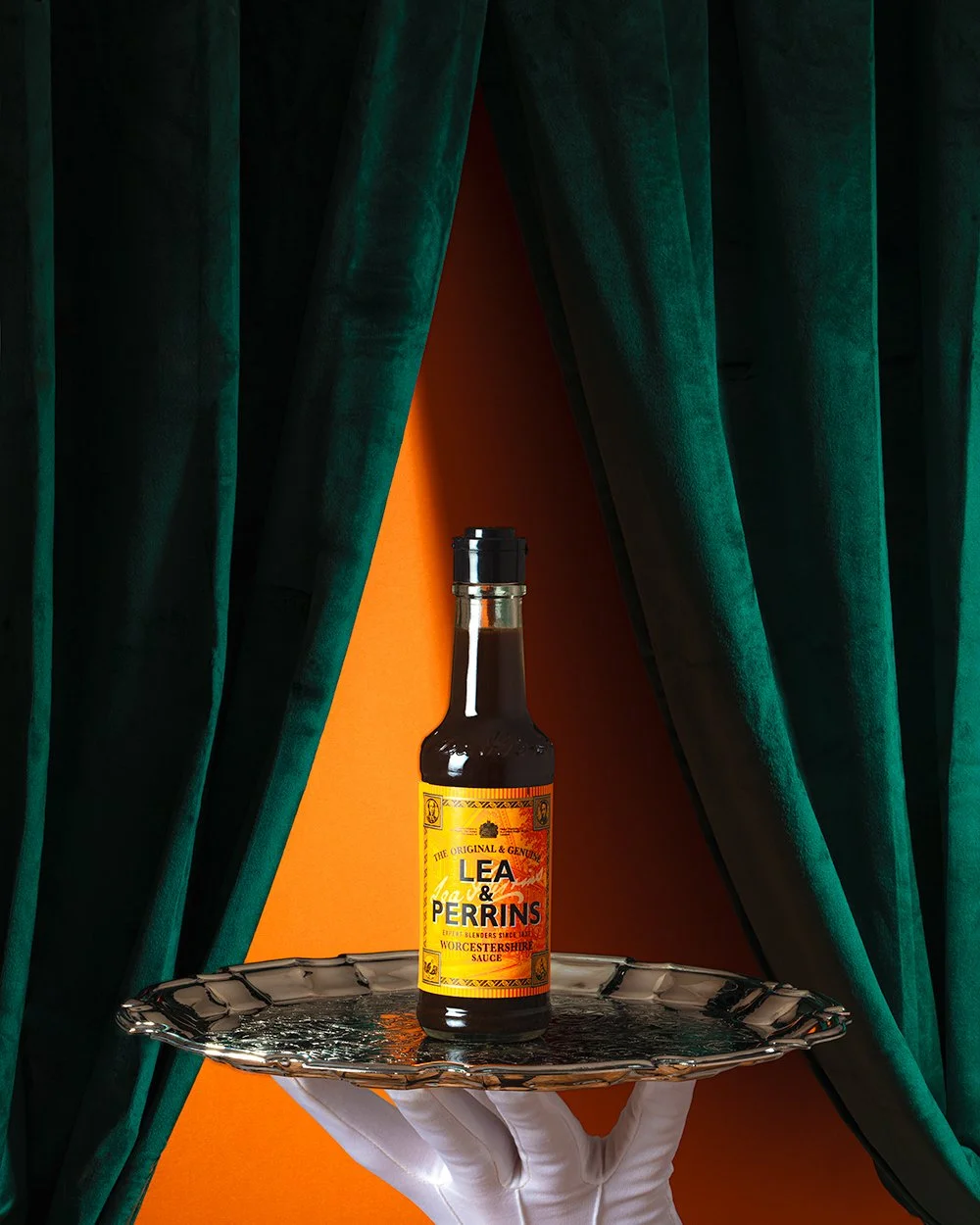 Lea & Perrins bottle of Worcestershire sauce elegantly displayed on a exquisite silver tray with green velvet curtain background and vibrant orange wall. 
