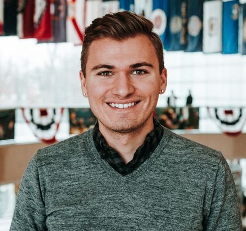A young man with short brown hair, wearing a grey sweater over a black collared shirt, smiling at the camera in a decorated indoor setting.