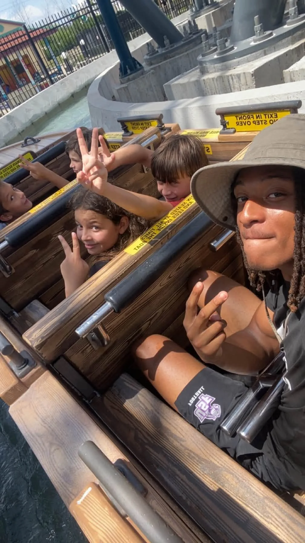 People sitting in a log-looking boat on water at an amusement park, with some making peace signs and smiling. The setting is sunny, with a metal fence and amusement park rides visible in the background.