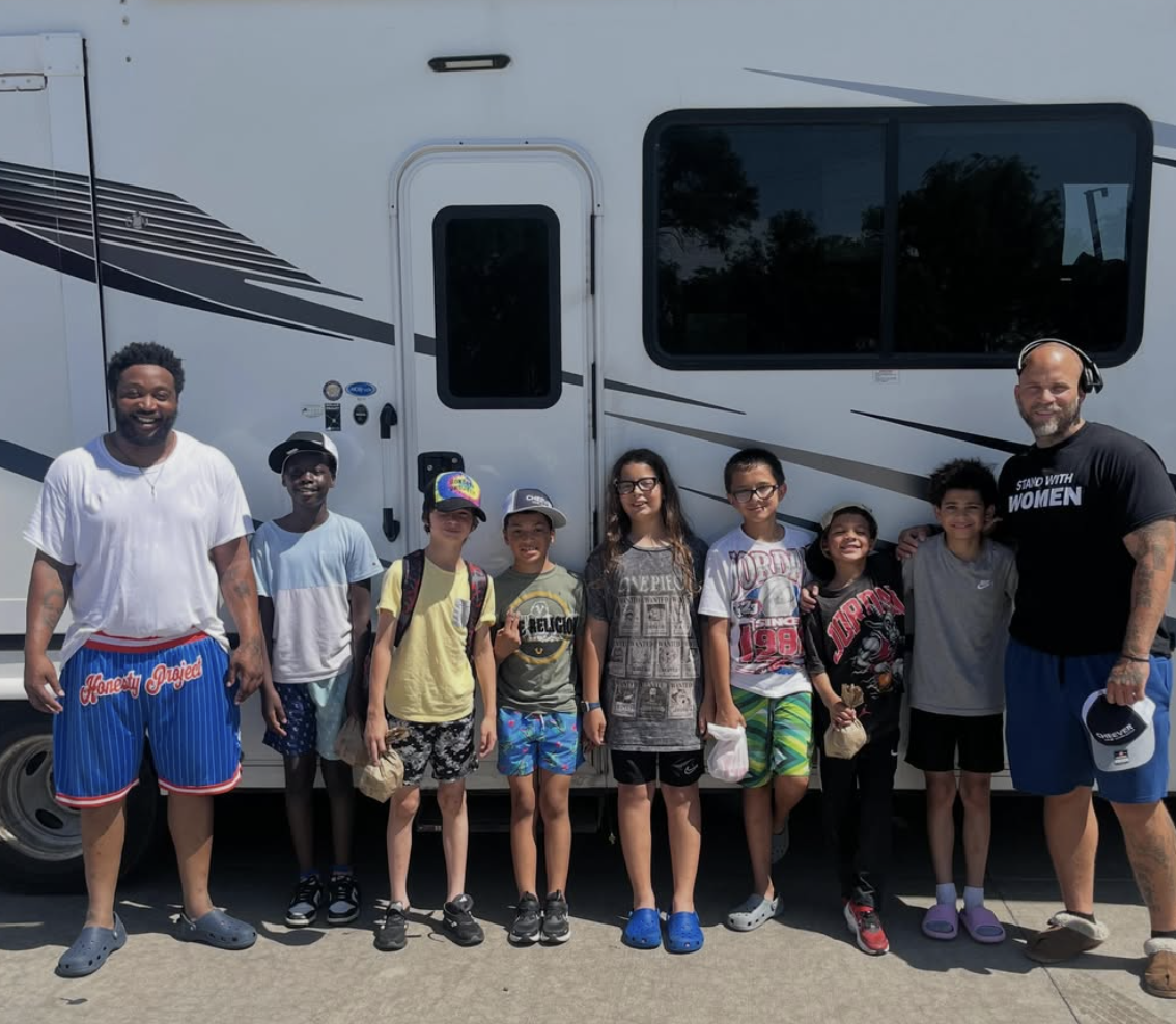 Group of children and two adults standing in front of an RV or camper, smiling and posing for a photo during daytime.