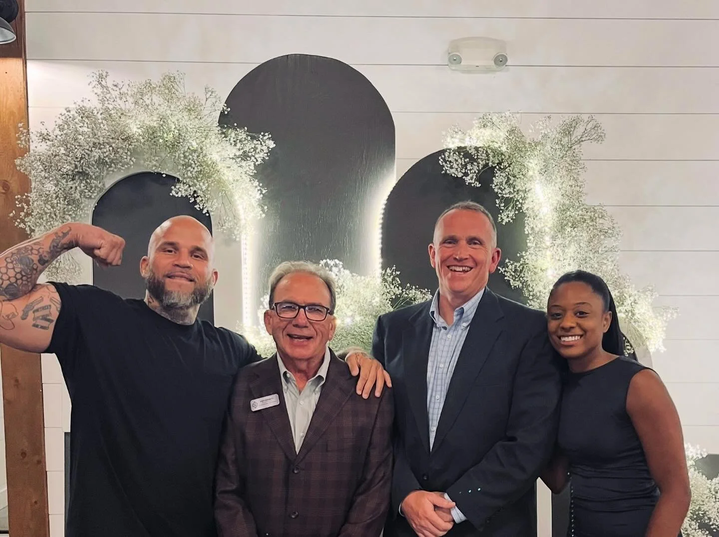 Four people standing together at an indoor event, smiling for the camera. The background features decorative elements with black circular shapes and white floral arrangements.