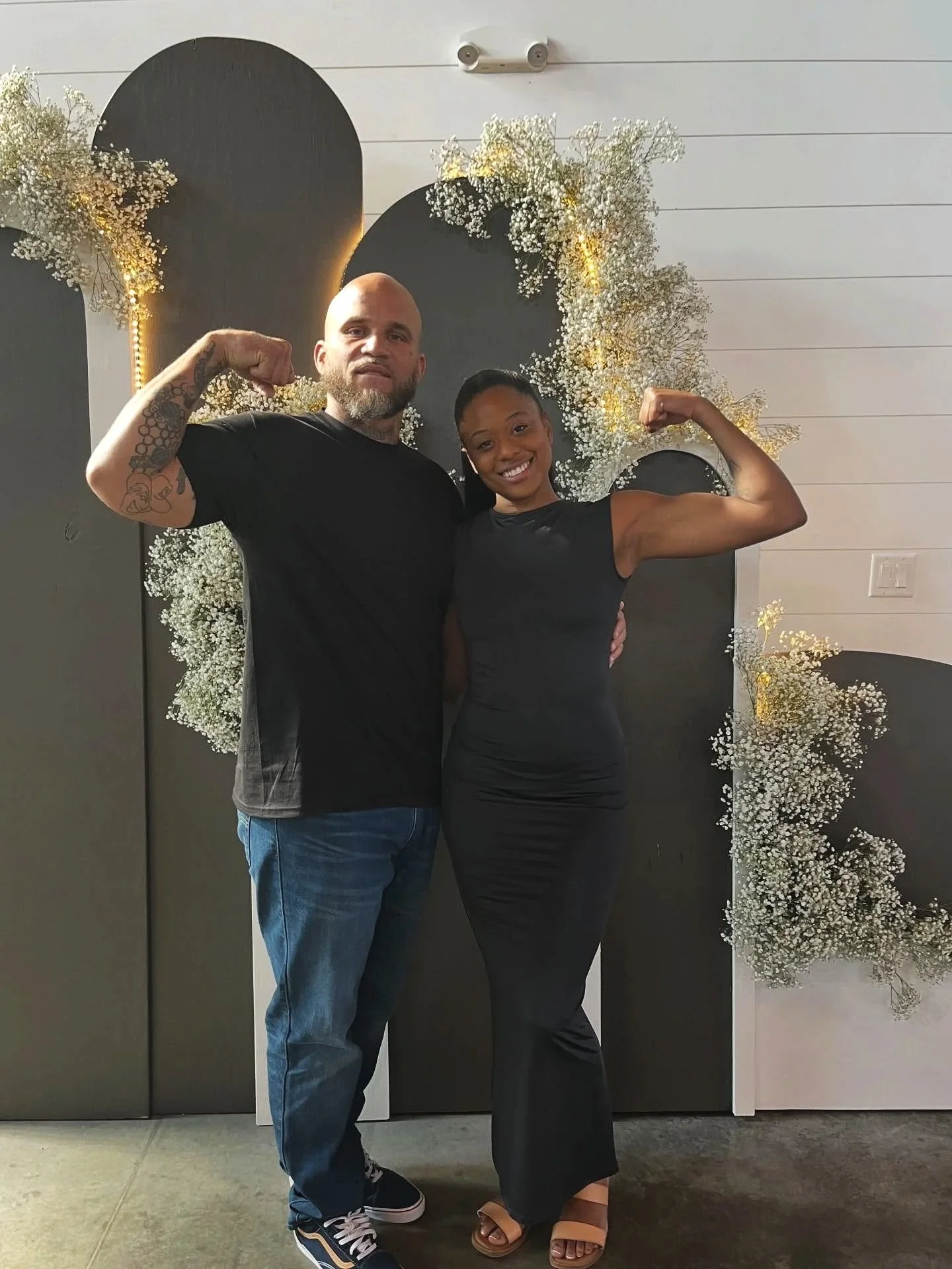 A man and woman posing with flexed biceps in front of a decorative backdrop with white flowers and black panels.