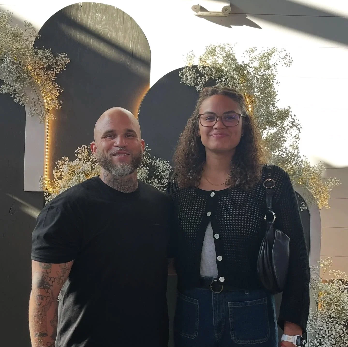 A man with a beard and tattoos on his arm stands next to a woman with curly hair and glasses; both are smiling, standing in front of decorative black circles with white flowers and warm lighting.