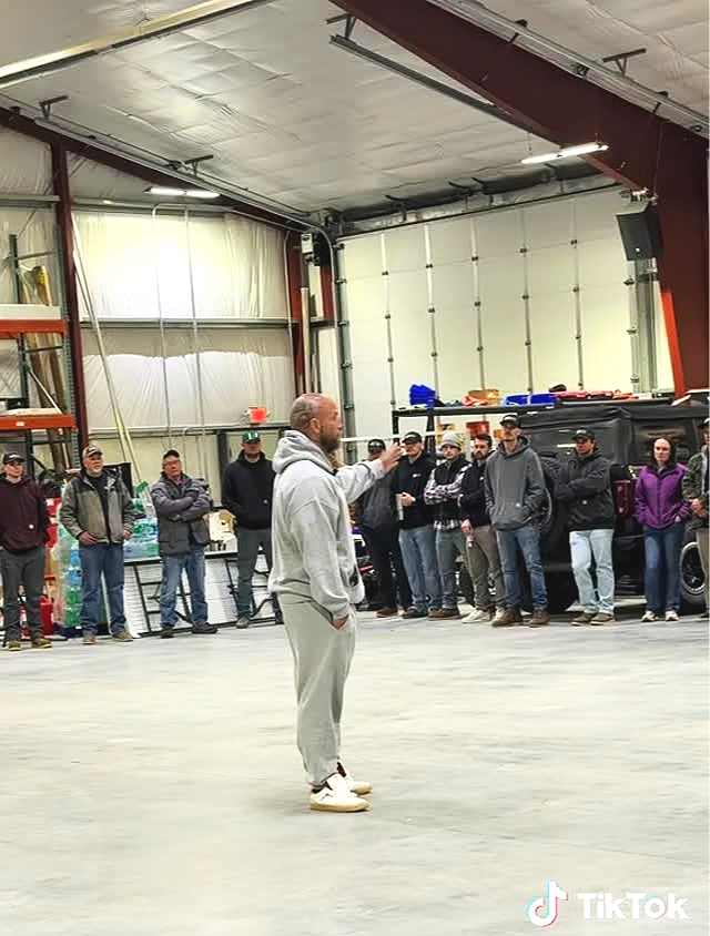 A man in a gray hoodie and sweatpants standing in the center of a large garage or warehouse, speaking to a group of people who are listening attentively, with vehicles and industrial shelving in the background.