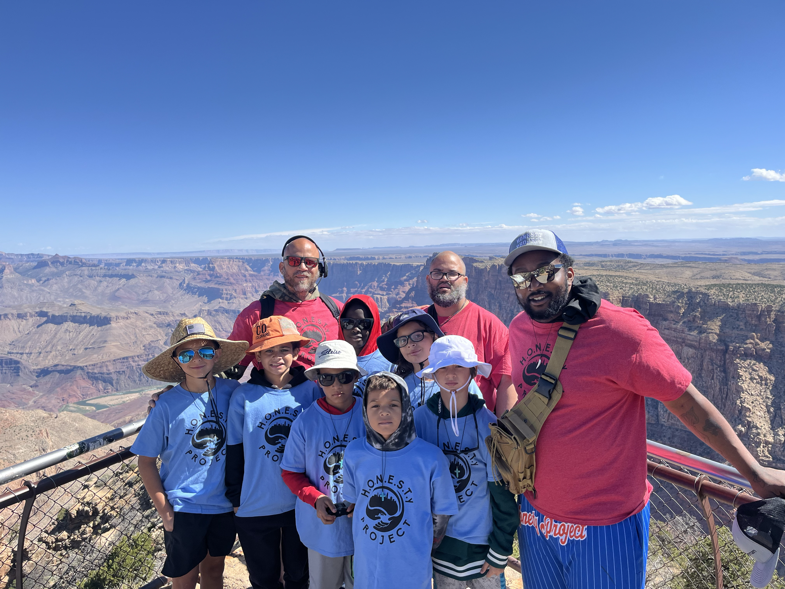 A group of children and adults standing together at the Grand Canyon with a scenic view of the canyon in the background, wearing casual clothes, some with sunglasses and hats, and smiling for the photo.
