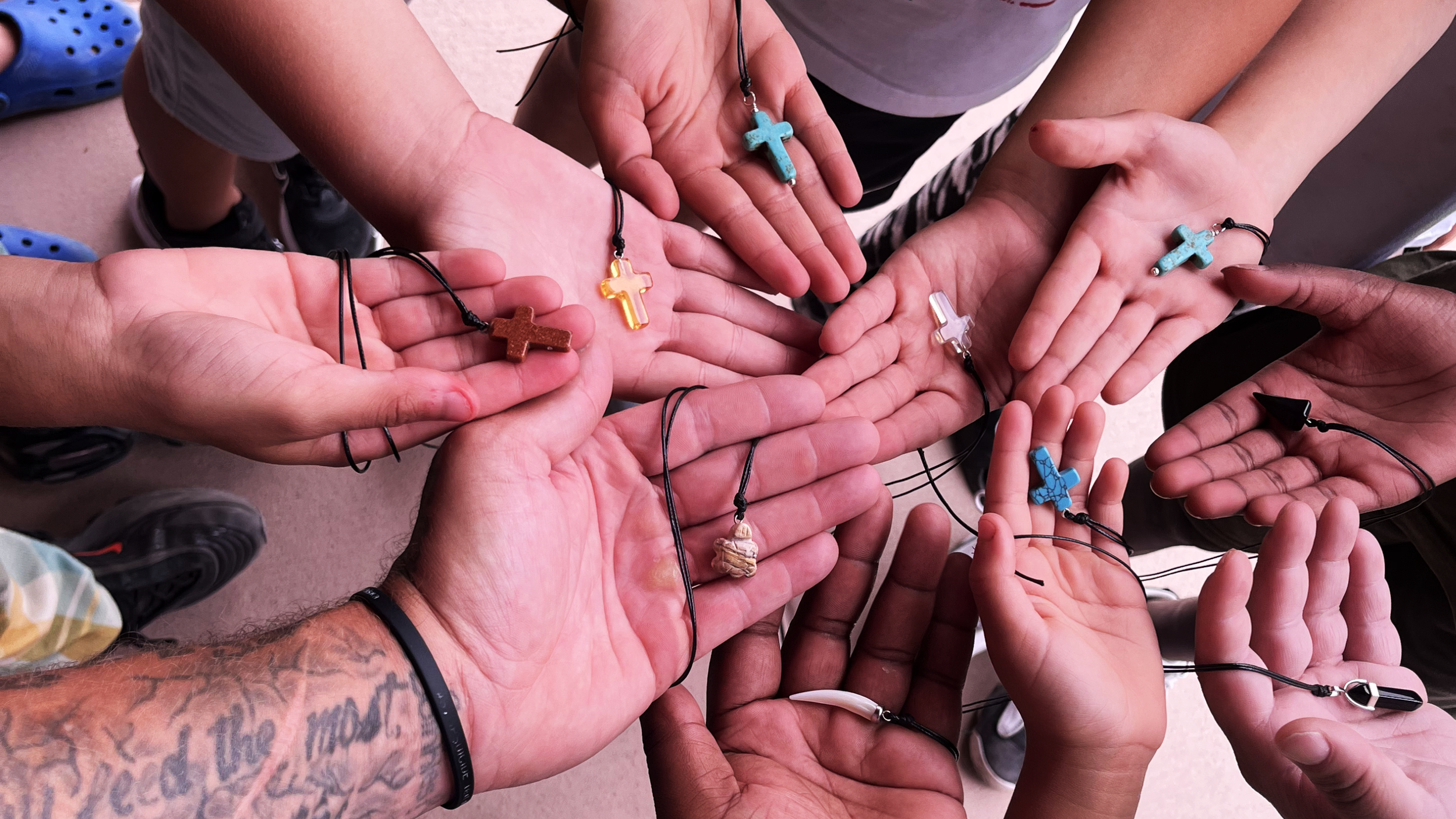 Group of people holding out their hands, each wearing a necklace with a cross pendant in various colors and styles.