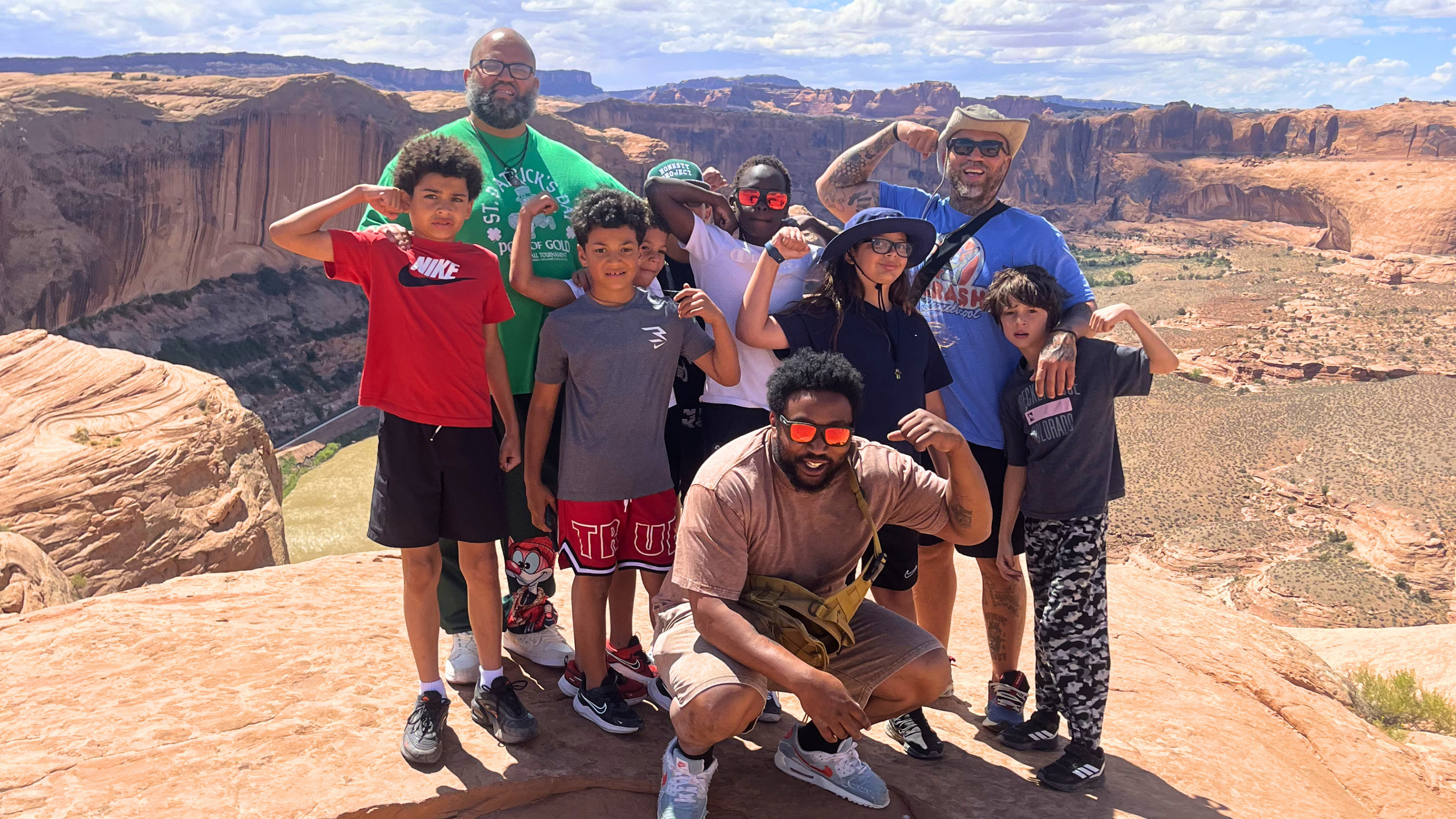 A group of people, including children and adults, posing for a photo on a rocky ledge in a desert canyon with cliffs and mesas in the background under a partly cloudy sky.