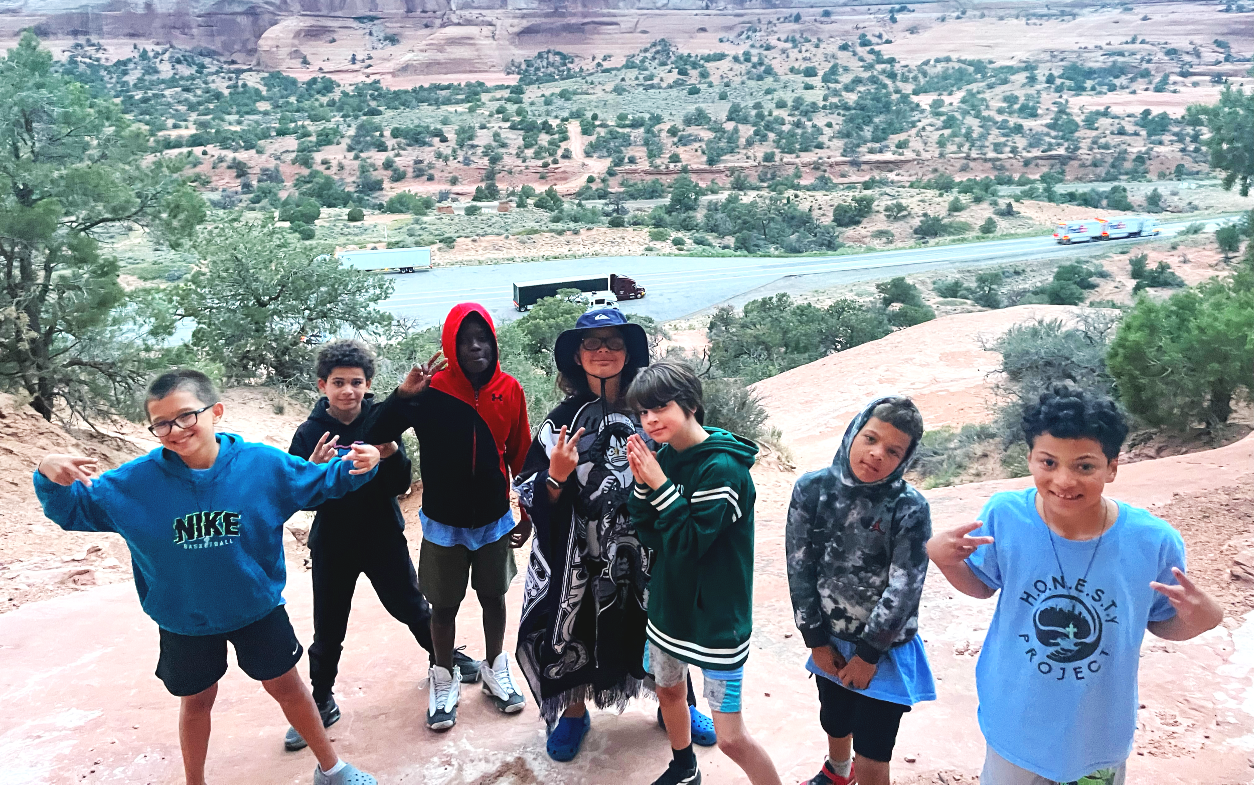 Group of seven children standing on a dirt trail in a desert landscape with red rock formations and sparse vegetation, some making peace signs and smiling.