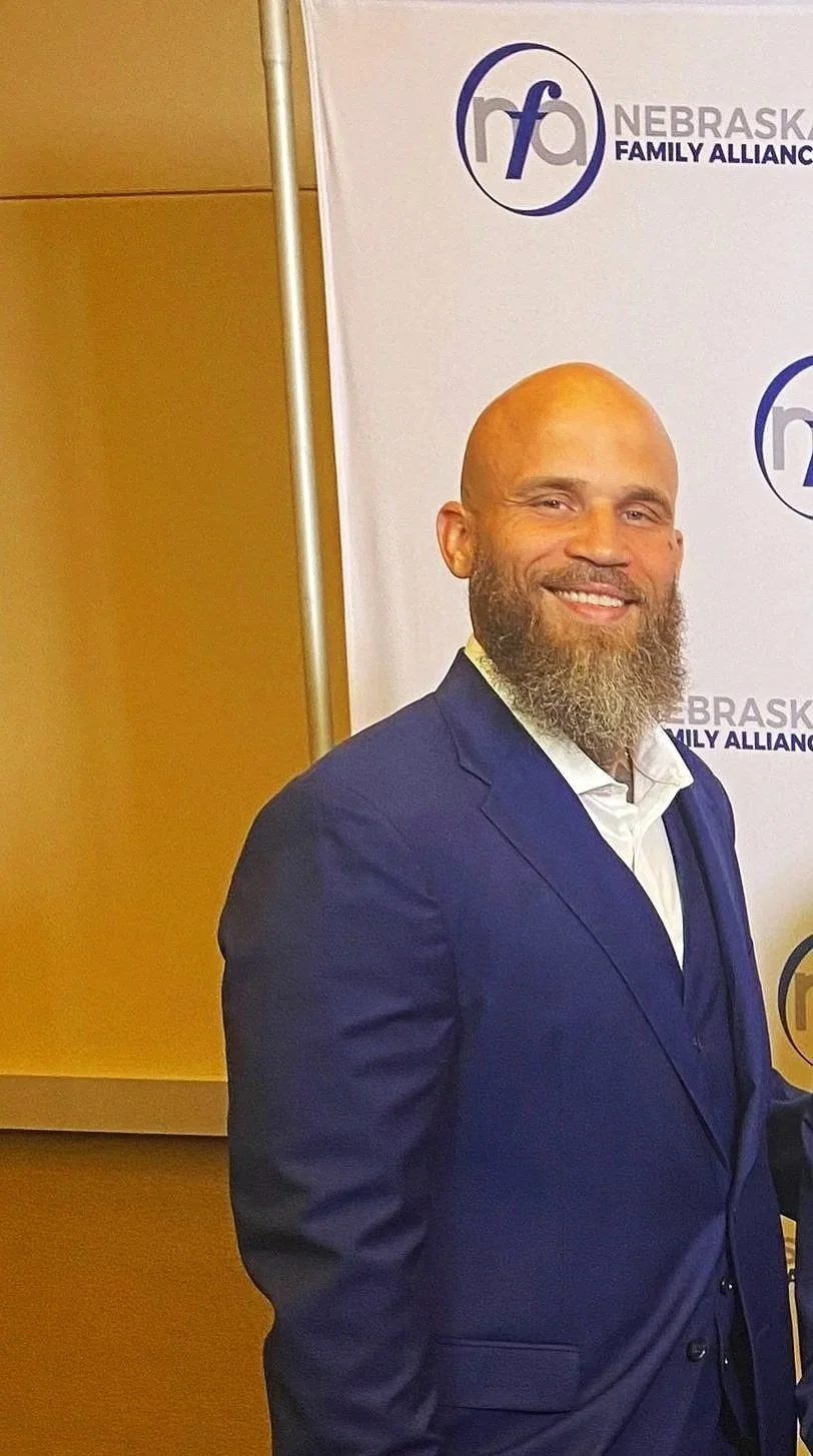 A man with a bald head and a beard, wearing a navy blue suit and white shirt, standing in front of a Nebraska Family Alliance banner with a logo and text.