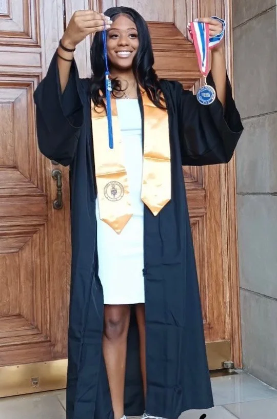 Woman in graduation gown holding medal and honor cords, smiling, standing in front of wooden door.