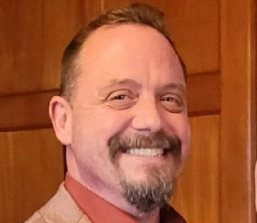 A smiling man with a beard and mustache, wearing a tan jacket over a red shirt, standing in front of wooden paneling.