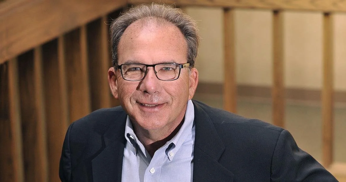 A middle-aged man with glasses, wearing a dark blazer over a light shirt, smiling at the camera against a background of wooden structure.