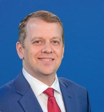 A man in a blue suit, white shirt, and red tie smiling against a blue background.