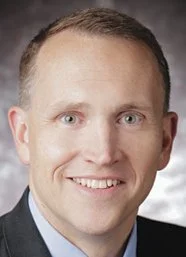 A professional headshot of a man with short light brown hair, blue eyes, and a friendly smile, wearing a dark suit and light shirt.