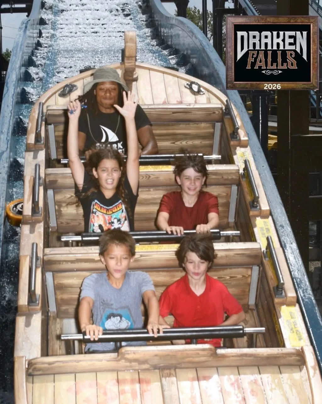 Five children on a wooden water ride at an amusement park, with water splashing around them, and a sign reading 'Draken Falls 2026' in the background.