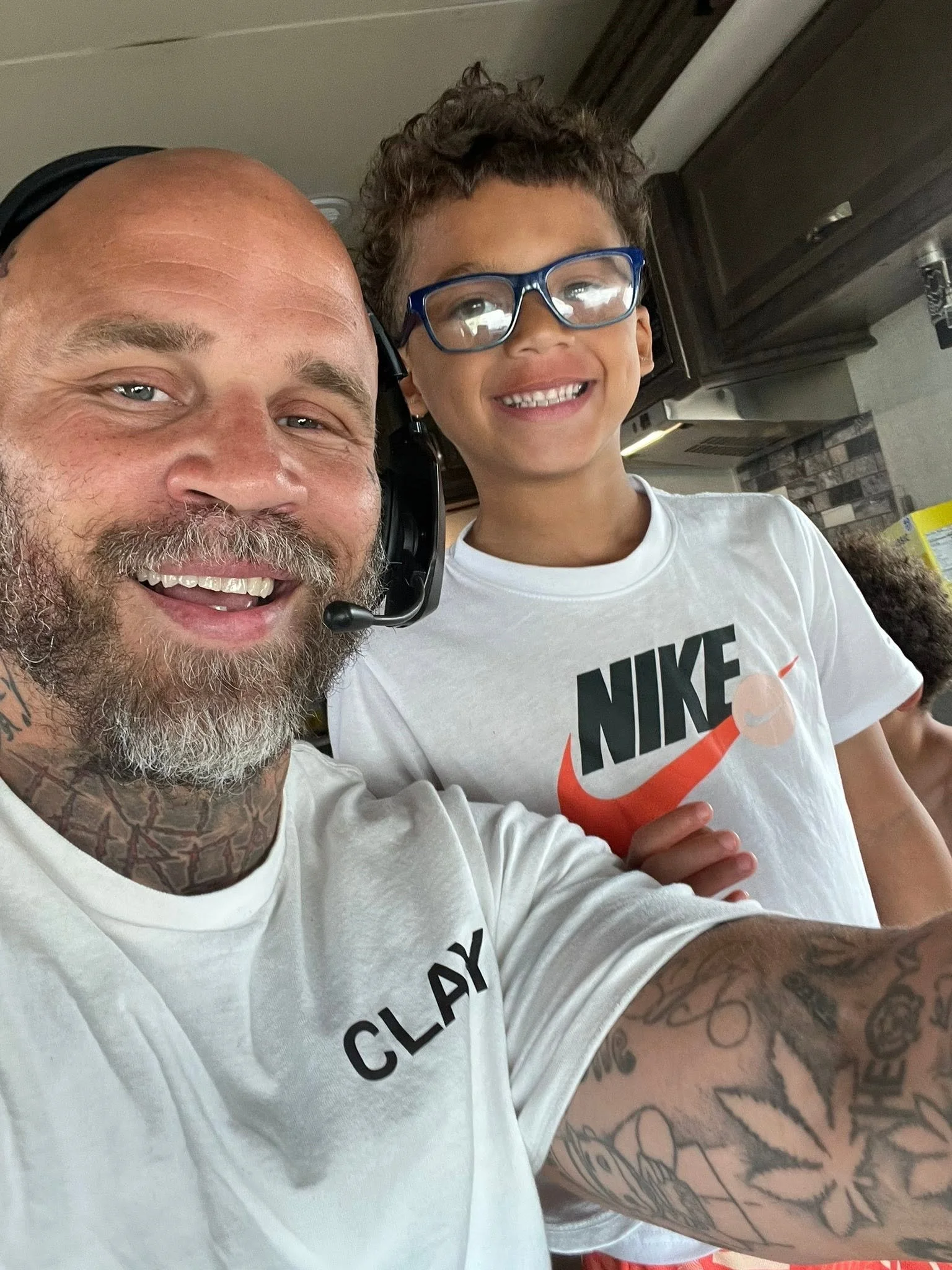 A man with a beard and tattoos on his arm, wearing a white shirt and headphones, smiling, taking a selfie with a young boy with curly hair and glasses, smiling, wearing a white Nike t-shirt.